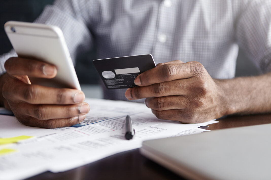 Modern technology and online shopping. African man in shirt paying for goods on internet using credit card and cell phone. Close-up of human hands, holding plastic card and mobile. Selective focus